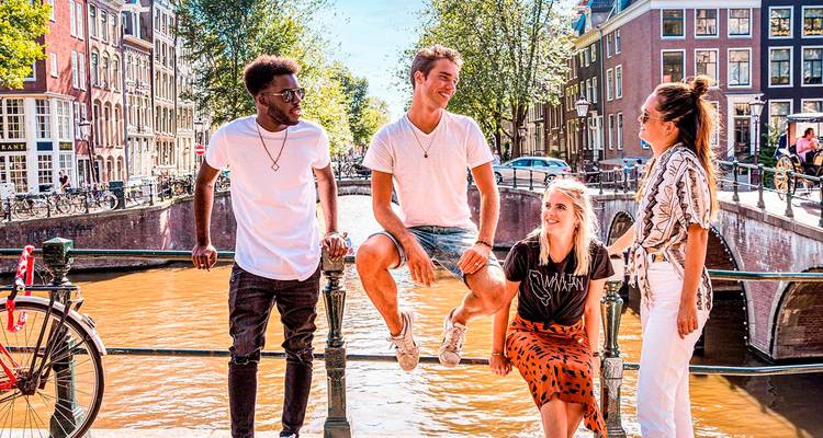 Four friends relax on a canal bridge under leafy trees with Amsterdam’s gabled houses lining the water.