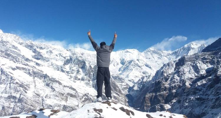 Person, die auf einem Schneegipfel steht mit erhobenen Händen, umgeben von Bergen.