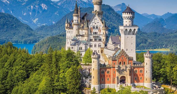 El castillo de cuento de hadas Neuschwanstein se alza sobre un bosque alpino con un lago azul y montañas escarpadas detrás.