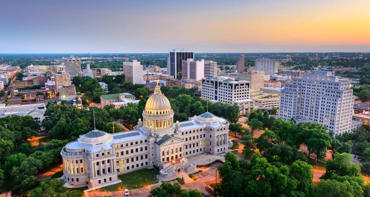 A cityscape at sunset with a capitol building and surrounding greenery.