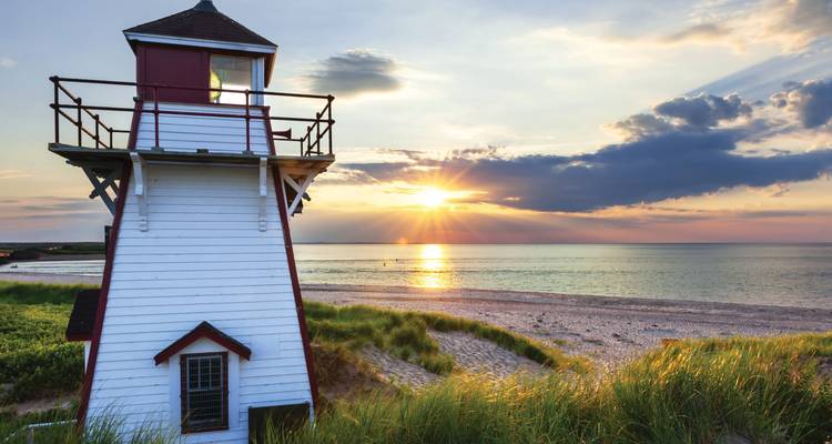 Un phare en bois côtier se dresse face à un coucher de soleil éclatant au-dessus d'une plage de sable tranquille et de la mer.