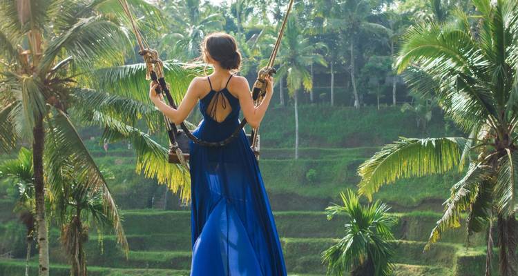 Frau in einem fließenden blauen Kleid auf einer Schaukel mit üppigem Grün und Reisfeldern.