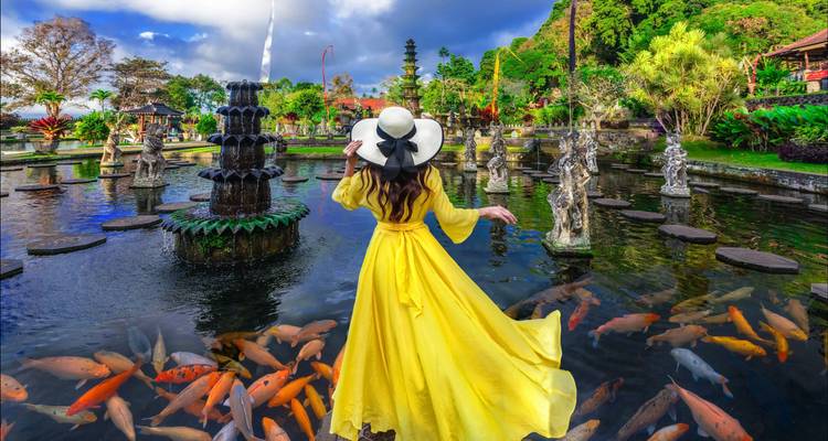 Frau in einem auffälligen gelben Kleid, die in einem Teich steht, umgeben von kunstvollen Statuen und Koi-Fischen.