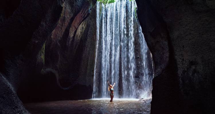 Frau steht fröhlich unter einem Wasserfall in einer natürlichen Höhlenumgebung.