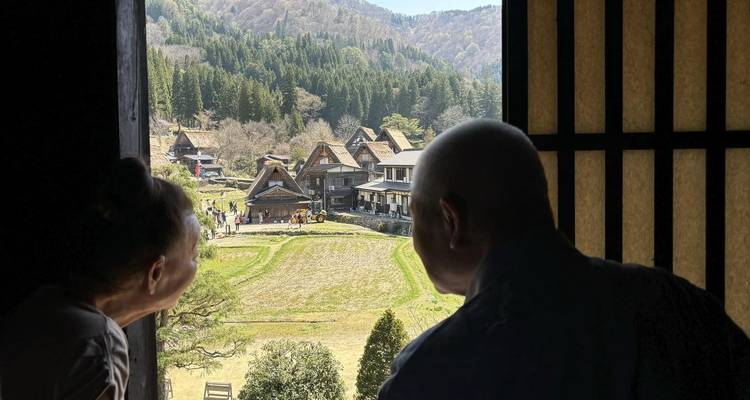 Dos viajeros asoman la cabeza por una ventana de madera tradicional hacia las casas de granja con techo de paja de la aldea de Shirakawago, rodeadas de bosques.