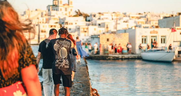 Young travellers walk along a stone pier beside whitewashed Cycladic buildings at golden hour on a Greek island harbour.