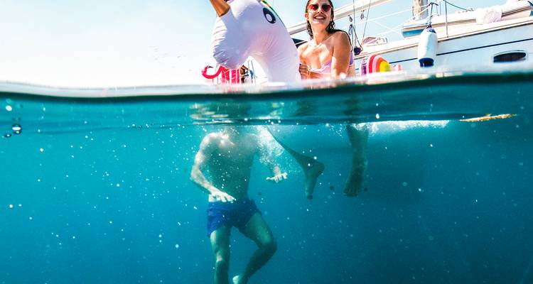Amigos juguetones disfrutan de un flotador de unicornio mientras nadan bajo un velero en aguas azules cristalinas.