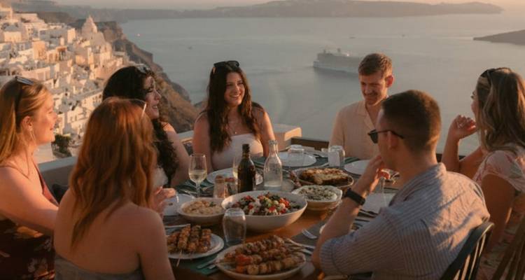 Gruppe von Freunden, die bei Sonnenuntergang ein Abendessen auf einer Terrasse mit Blick auf die Caldera von Santorin geniessen.