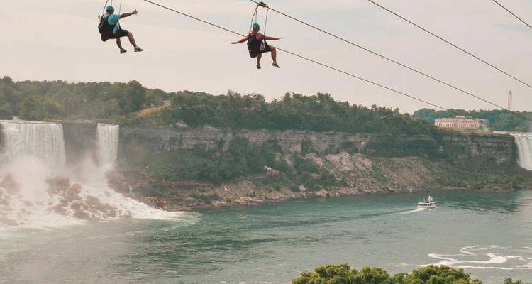 Zwei Abenteuerlustige gleiten auf einer Seilrutsche über den Niagara River, während der Nebel der Horseshoe Falls hinter ihnen aufsteigt.