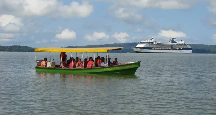 Petit bateau à moteur vert transportant des touristes sur un grand lac avec un grand navire de croisière ancré en arrière-plan sous un ciel bleu et des nuages cotonneux.