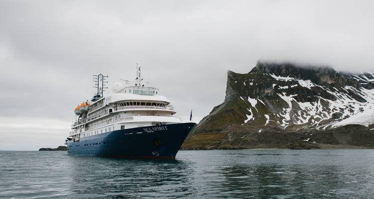 Cruise ship sailing in front of rugged, snow-capped terrain.