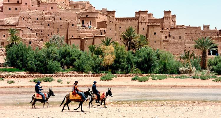 Menschen auf Eseln reiten an traditionellen Lehmgebäuden vorbei.
