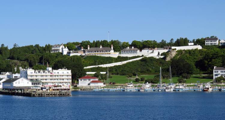 Vue du front de mer de la marina de l'île Mackinac, des fortifications historiques et de la colline verdoyante sous un ciel dégagé.