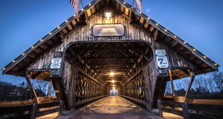 Pont couvert en bois illuminé par des lumières chaudes au crépuscule, perspective qui s'étend le long de sa passerelle en planches.