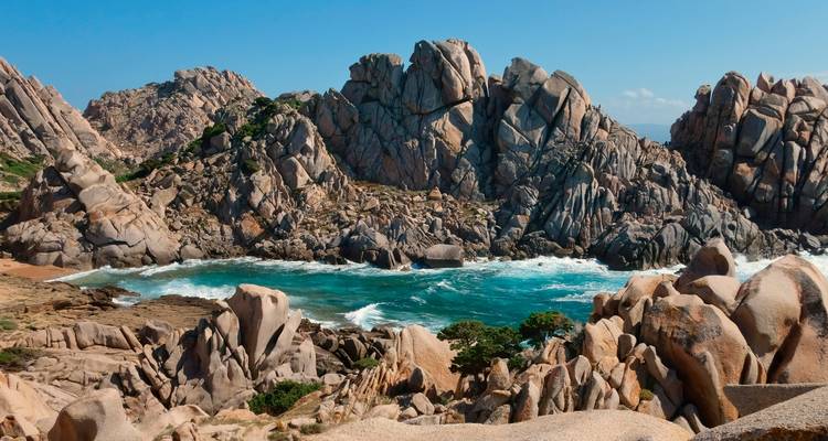 Dramatische Granitfelsen und türkisfarbene Bucht mit brandenden Wellen an einem sonnigen Tag.