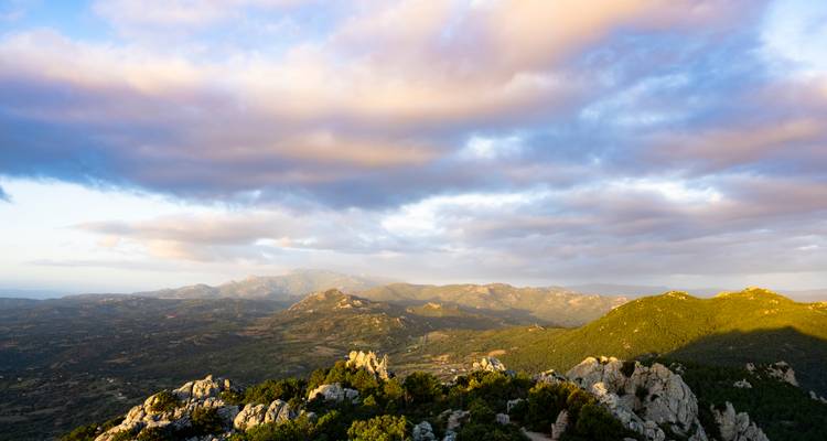 Hügeliges Berglandschaft unter farbenfrohen Abendrwolken, von einem felsigen Bergrücken aus betrachtet.