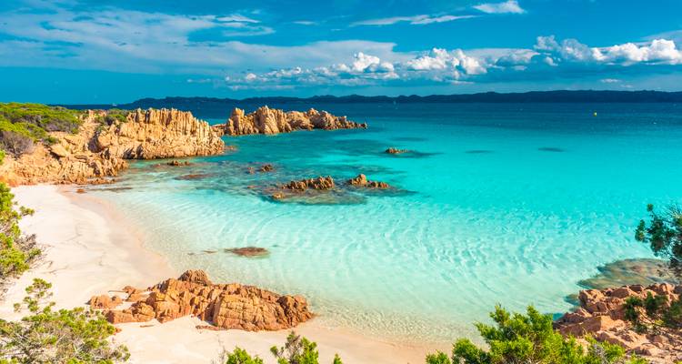 Unberührter Strand mit weißem Sand, leuchtendem türkisfarbenem Wasser und zerklüfteten Felsformationen unter einem strahlend blauen Himmel.