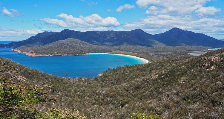 Vaste panorama de la baie Wineglass Bay avec son croissant parfait de sable blanc et d'eau turquoise encadré par des montagnes boisées