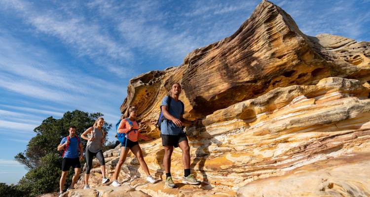Grupo activo de senderismo pasando por formaciones de arenisca estratificada bajo un cielo azul brillante.