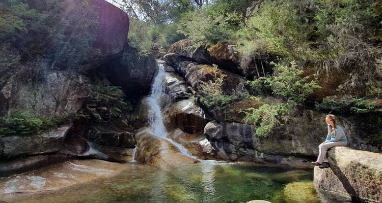 Persoon zittend bij een waterval omringd door rotsen en groen.