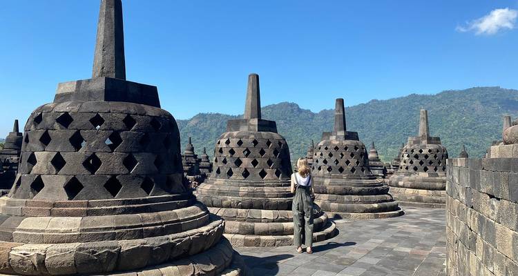 Viajero fotografiando las estupas con forma de campana del templo Borobudur contra cielos azules despejados y montañas distantes.