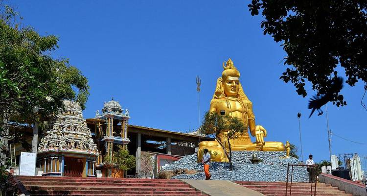Une grande statue dorée de Shiva est située sur un complexe de temples au sommet d'une colline, se détachant contre un ciel bleu dégagé.