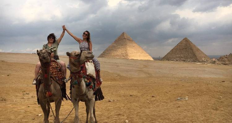 Dos personas en camellos con pirámides al fondo.