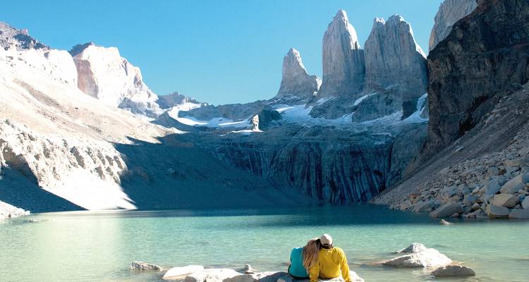 A couple relaxes beside a turquoise alpine lake with the jagged granite towers of Torres del Paine rising in the background under a bright blue sky.