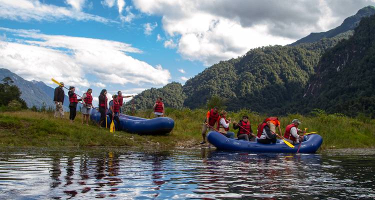 Reizigers met reddingsvesten laten blauwe rubberbootjes op een rustige rivier af, omgeven door weelderig groene bergen en een gedeeltelijk bewolkte hemel.