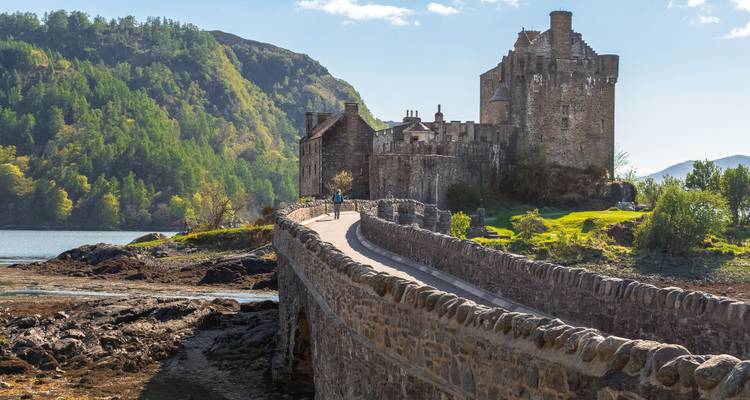 Un château sur une petite île relié par une passerelle, entouré d'eau et de collines.