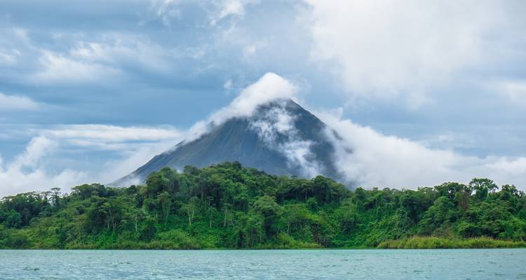 De met wolken bedekte vulkaan Arenal torent uit boven groen regenwoud en een blauw meer.