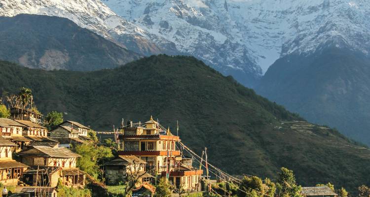 Bergdorf mit traditionellen Häusern auf einem grünen Hang gelegen, mit schneebedeckten Himalaya-Gipfeln im Hintergrund.