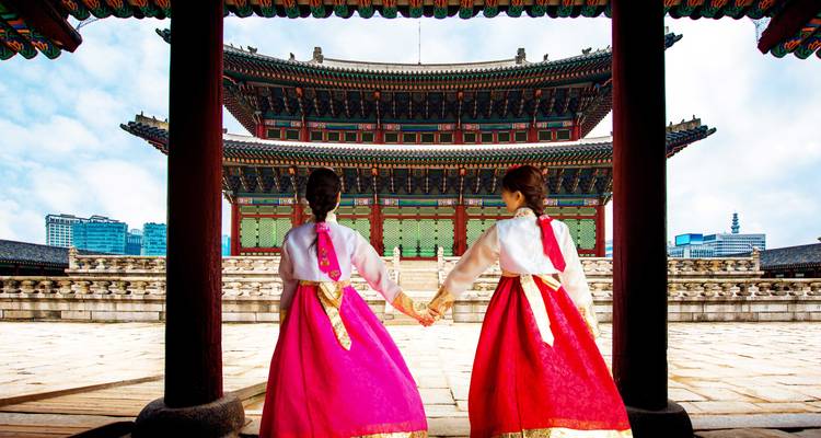 Dos mujeres con hanbok de colores brillantes se toman de las manos mientras contemplan una sala de palacio ornamentada.