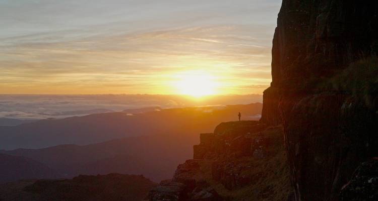 A person standing on a cliff overlooking a valley during sunset.