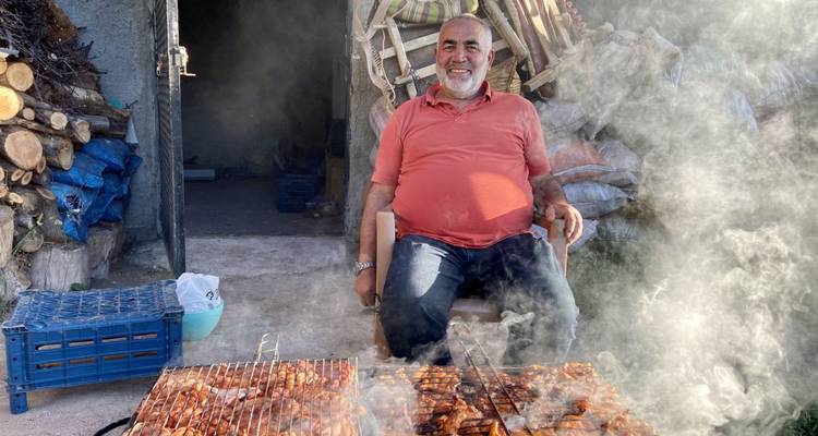 Mann, der neben einem Grill mit Rauch sitzt, umgeben von rustikaler Außenkulisse.
