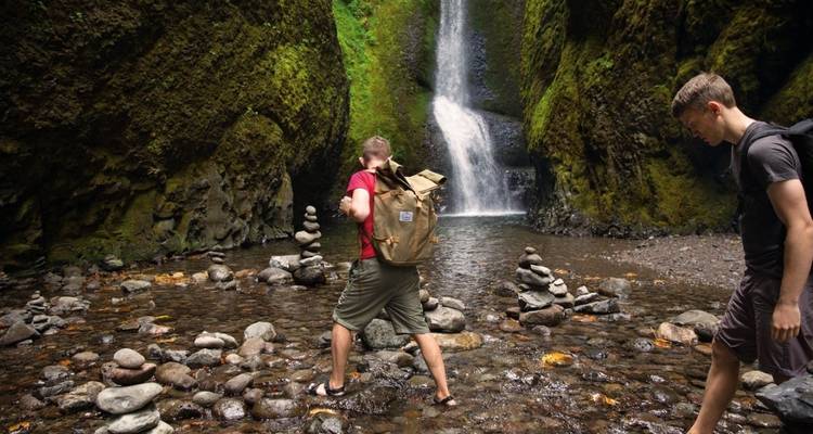 Zwei Wanderer überqueren einen seichten Bach mit einem Wasserfall.
