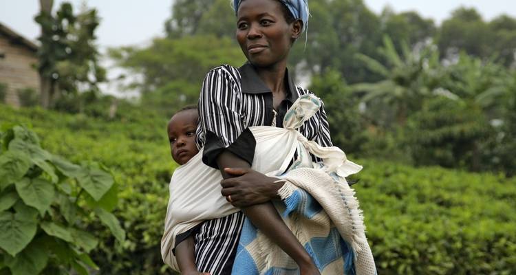 A woman carrying a baby on her back walking through greenery.