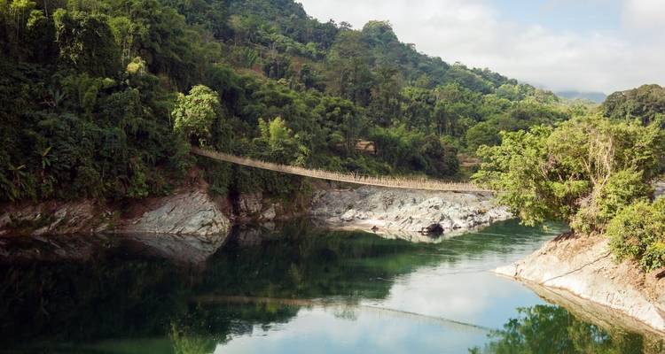 Eine Hängebrücke überspannt einen breiten Fluss mit üppigem Grün und Hügeln.