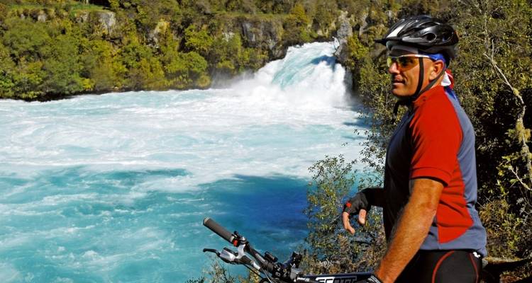 Cyclist standing by a river with a waterfall in the background.