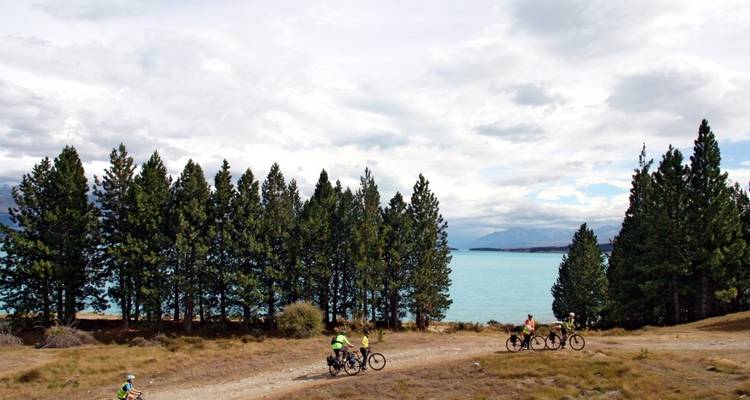 Cyclists on a trail by a turquoise lake with pine trees.