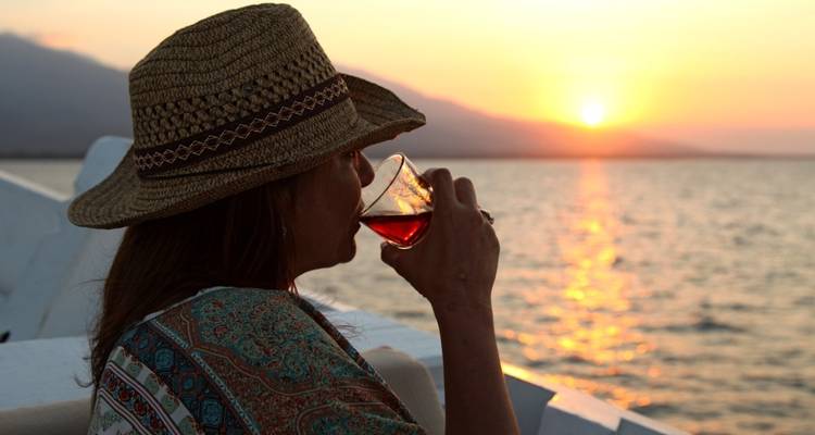 Vrouw met een hoed die een drankje drinkt terwijl ze naar de zonsondergang over het water kijkt.
