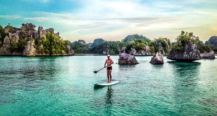 Persoon die paddleboardt in helder water omringd door rotsachtige eilanden.