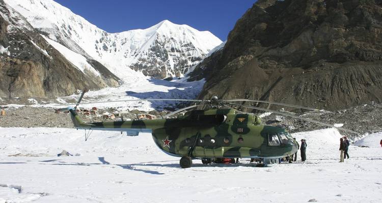 Military helicopter on a snowy field with mountains, people nearby working.