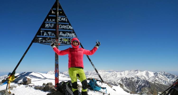 Person posiert auf einem Berggipfel mit einer Metallpyramiden-Skulptur.