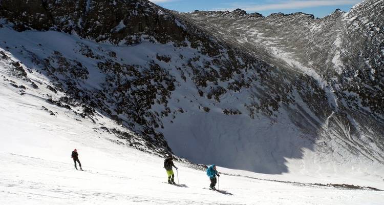 Drei Personen fahren Ski einen verschneiten Hang hinunter mit einem großen Berg im Hintergrund.