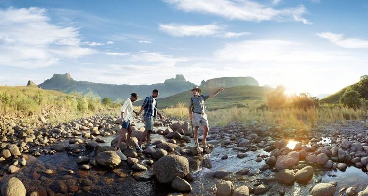 Three people crossing a rocky stream in a mountainous landscape.