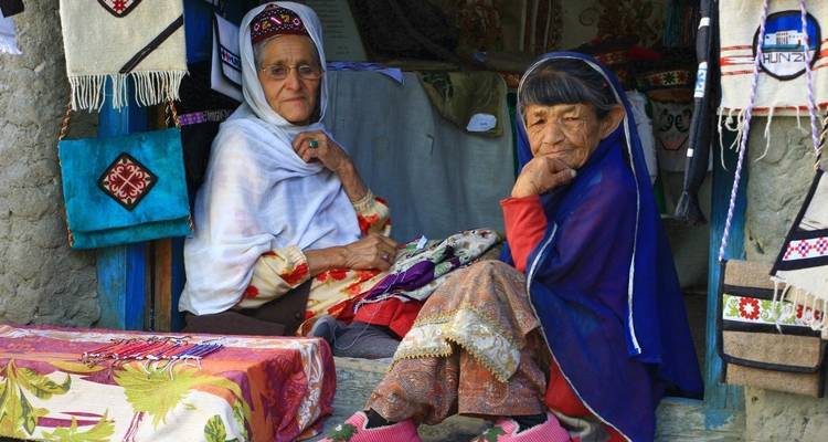 Older women sitting in front of a shop with textiles, engaging with viewers.