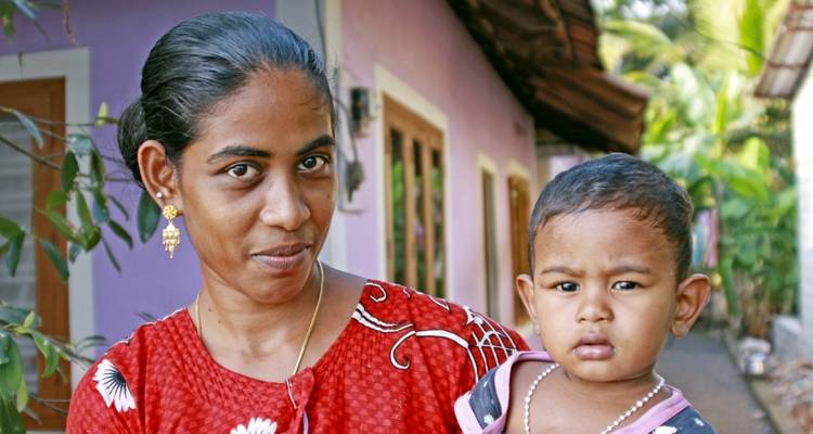 Une femme tenant un enfant, debout à l'extérieur avec des maisons en arrière-plan.