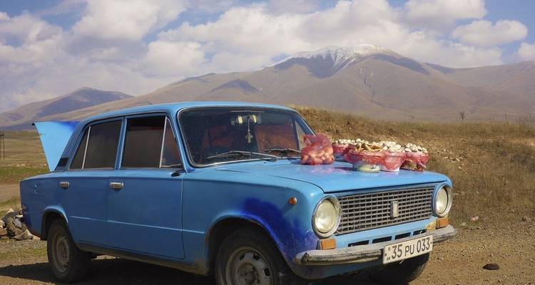 A vintage blue car with fruits and vegetables in front of a mountain backdrop.