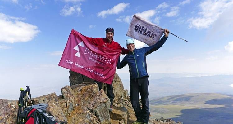 Two people holding flags on a rocky summit with a view of distant plains.